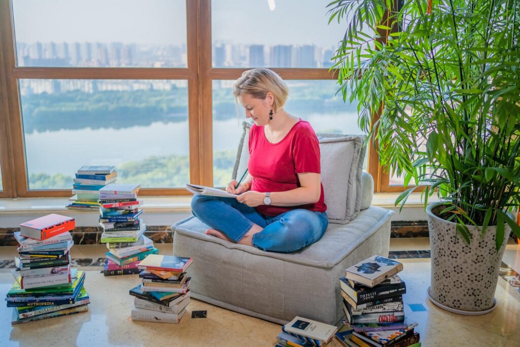 Vor Panoramafenster auf Sessel sitzend schreibt Tanja Wirnitzer in ein Heft, umgeben von einer großen grünen Pflanze und vielen Buchstapeln auf dem Boden.